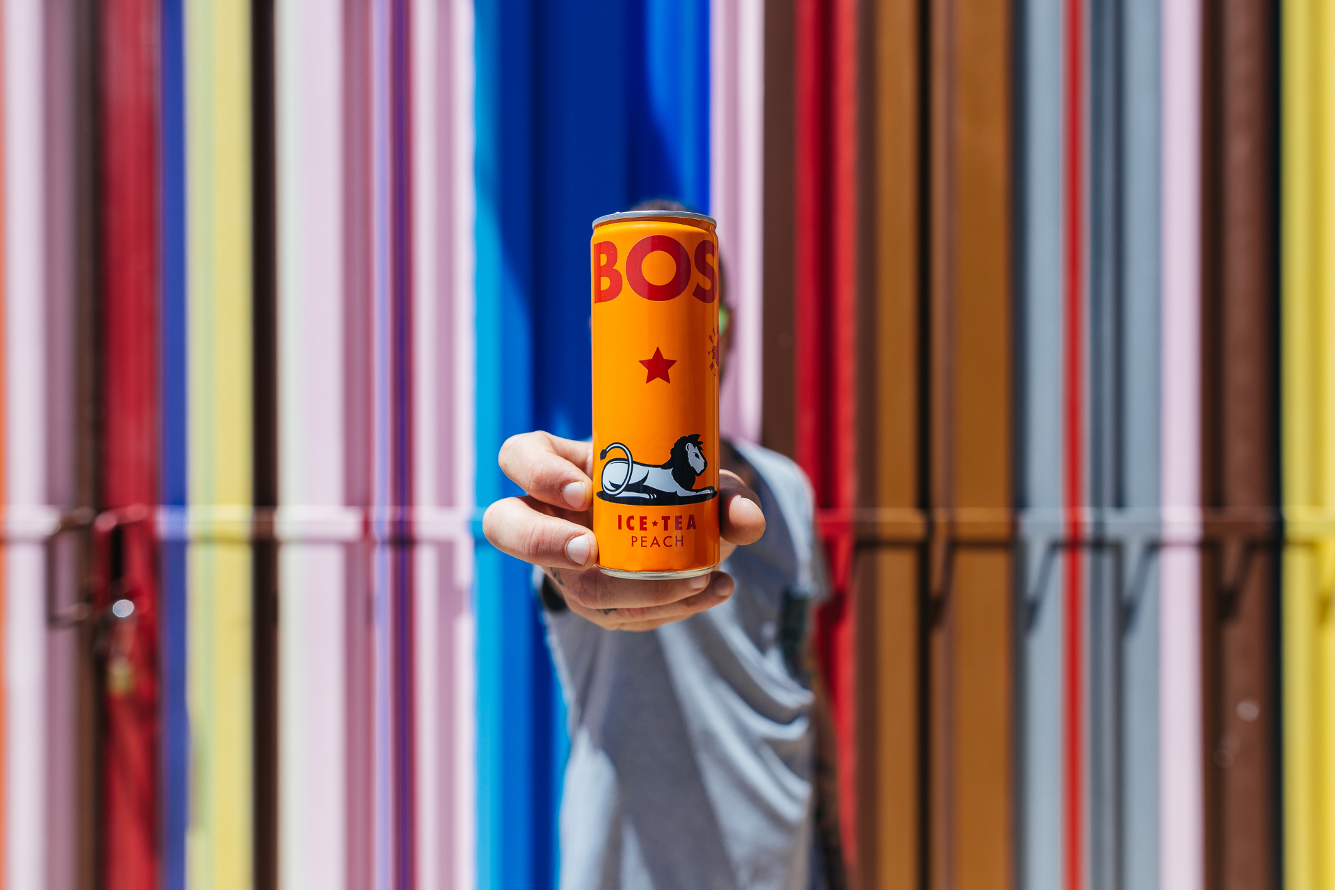 Hand holding a BOS Peach Ice Tea can in front of a colourful striped door