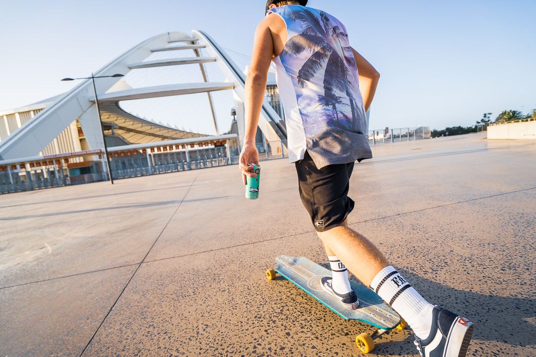 Skater carrying a BOS can walking towards a stadium, shot from behind