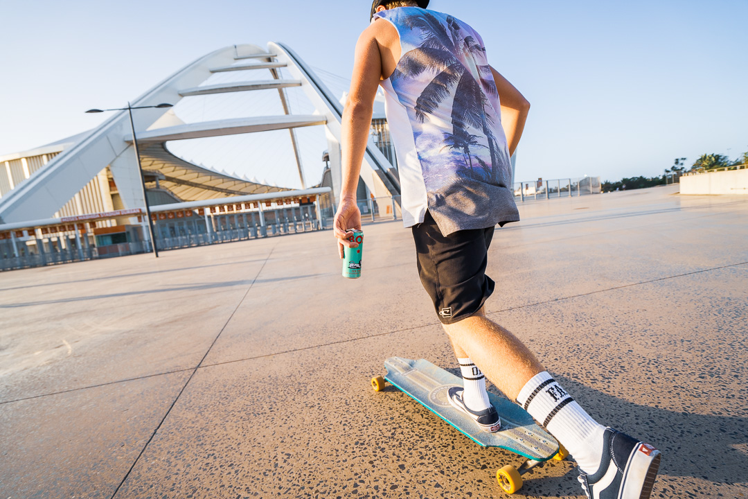 Skater carrying a BOS can walking towards a stadium, shot from behind